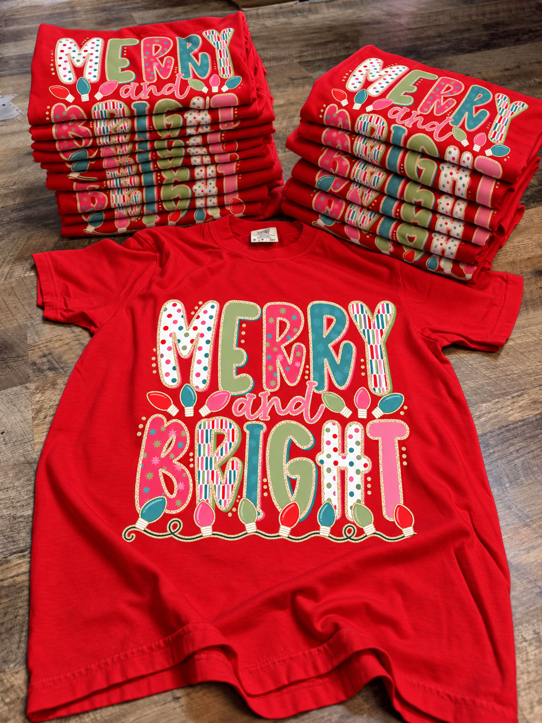 Merry and Bright red Christmas graphic tees stacked on rustic wood with colorful holiday lettering and lights