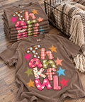 Colorful THANKFUL patchwork graphic on warm taupe-brown tee next to stacked tees and blanket