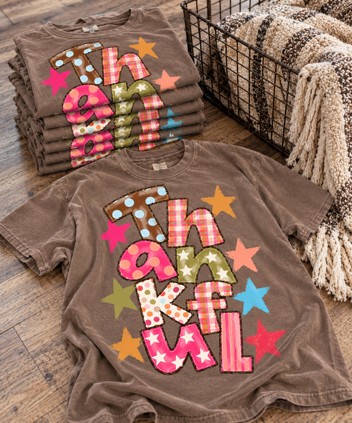 Colorful THANKFUL patchwork graphic on warm taupe-brown tee next to stacked tees and blanket